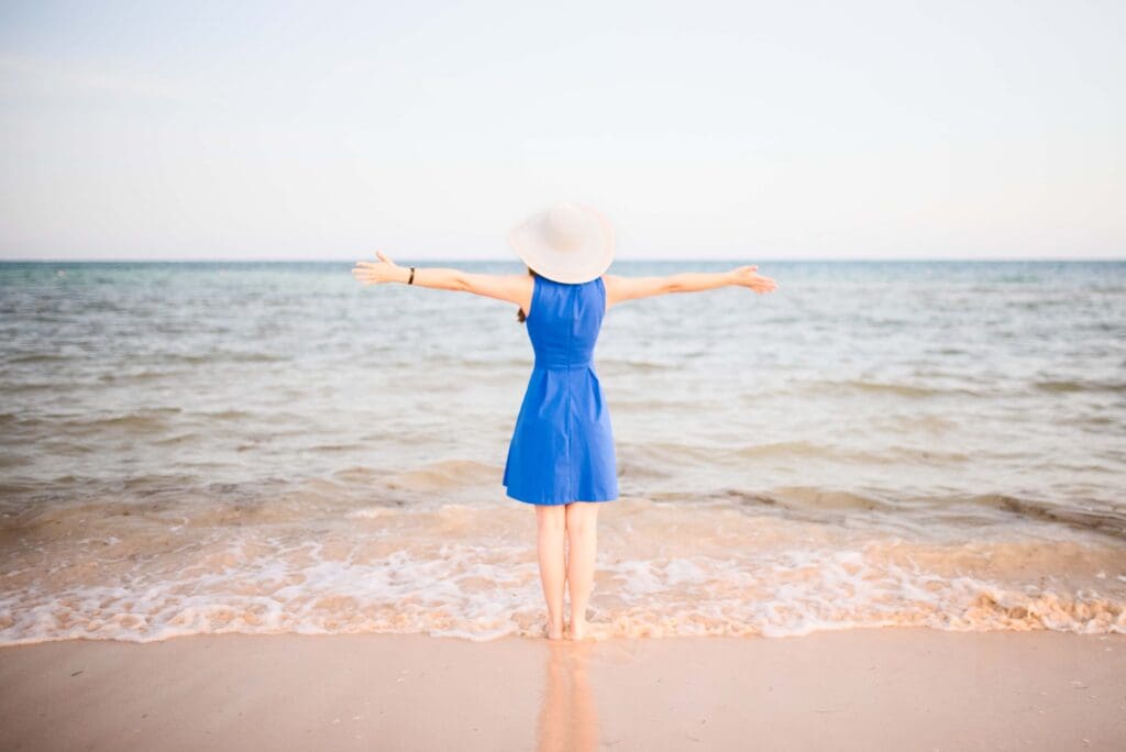 Woman in blue dress standing on beach with arms open toward ocean